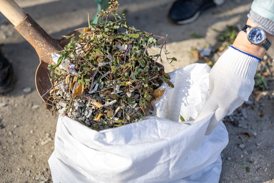 A close-up view of a gardener's hands wearing white gloves, collecting a small pile of mixed garden debris, including dried leaves, twigs, and small rocks, into a white outdoor storage bag. In the bac